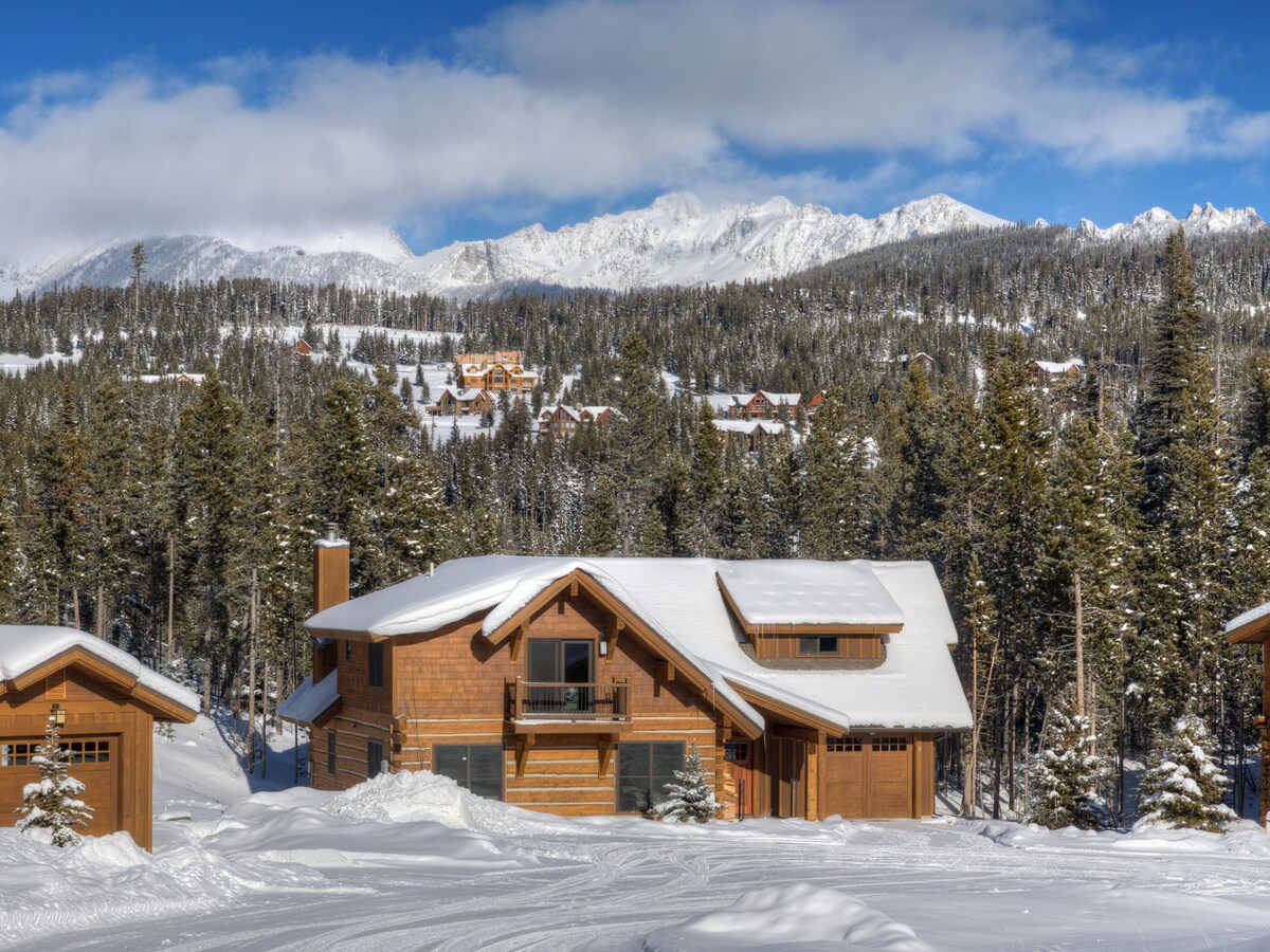 The exterior of a log cabin is positioned among snow-covered pines, with the mountains visible in the distance. The cabin features large windows, a sloped roof, and a driveway leading to the entrance, all set against a clear blue sky.