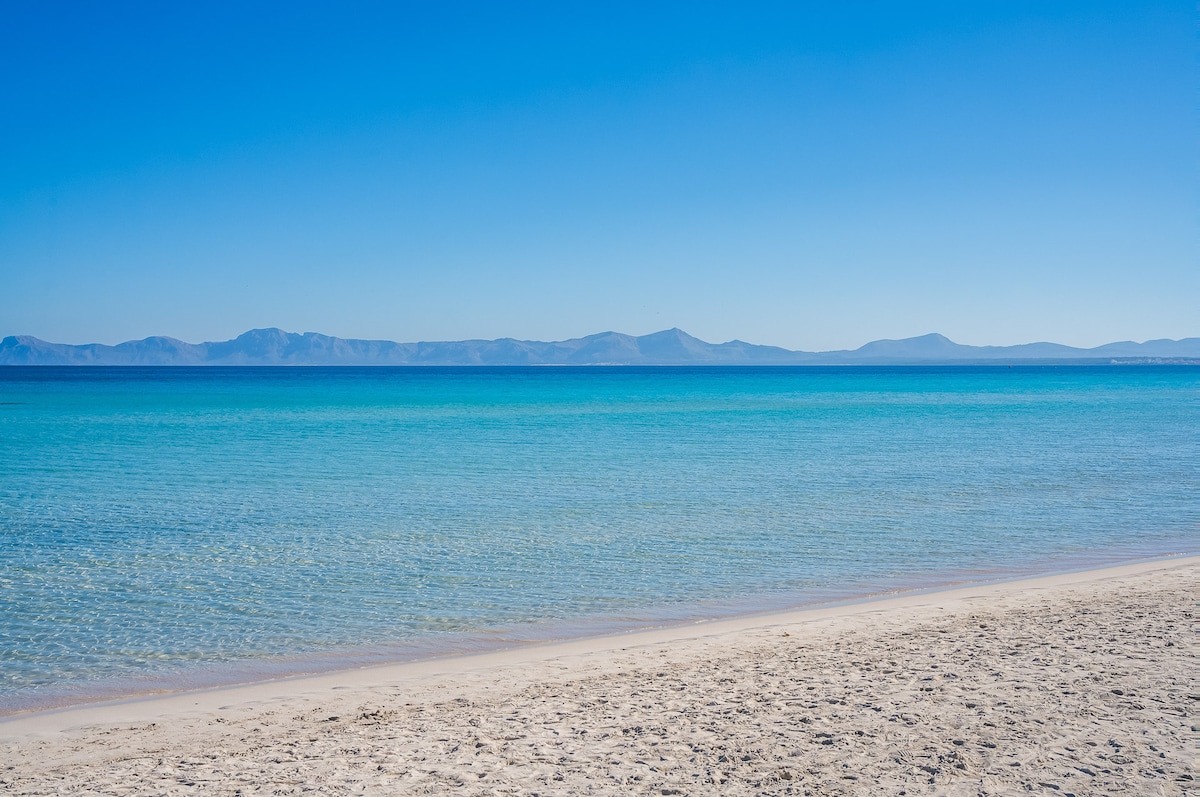 A serene beach view showcases soft white sand meeting calm turquoise waters. Gentle waves lap against the shore, while distant mountains fade into a clear blue sky. The tranquil scene highlights the natural beauty of the coastline, inviting relaxation and peaceful reflection.