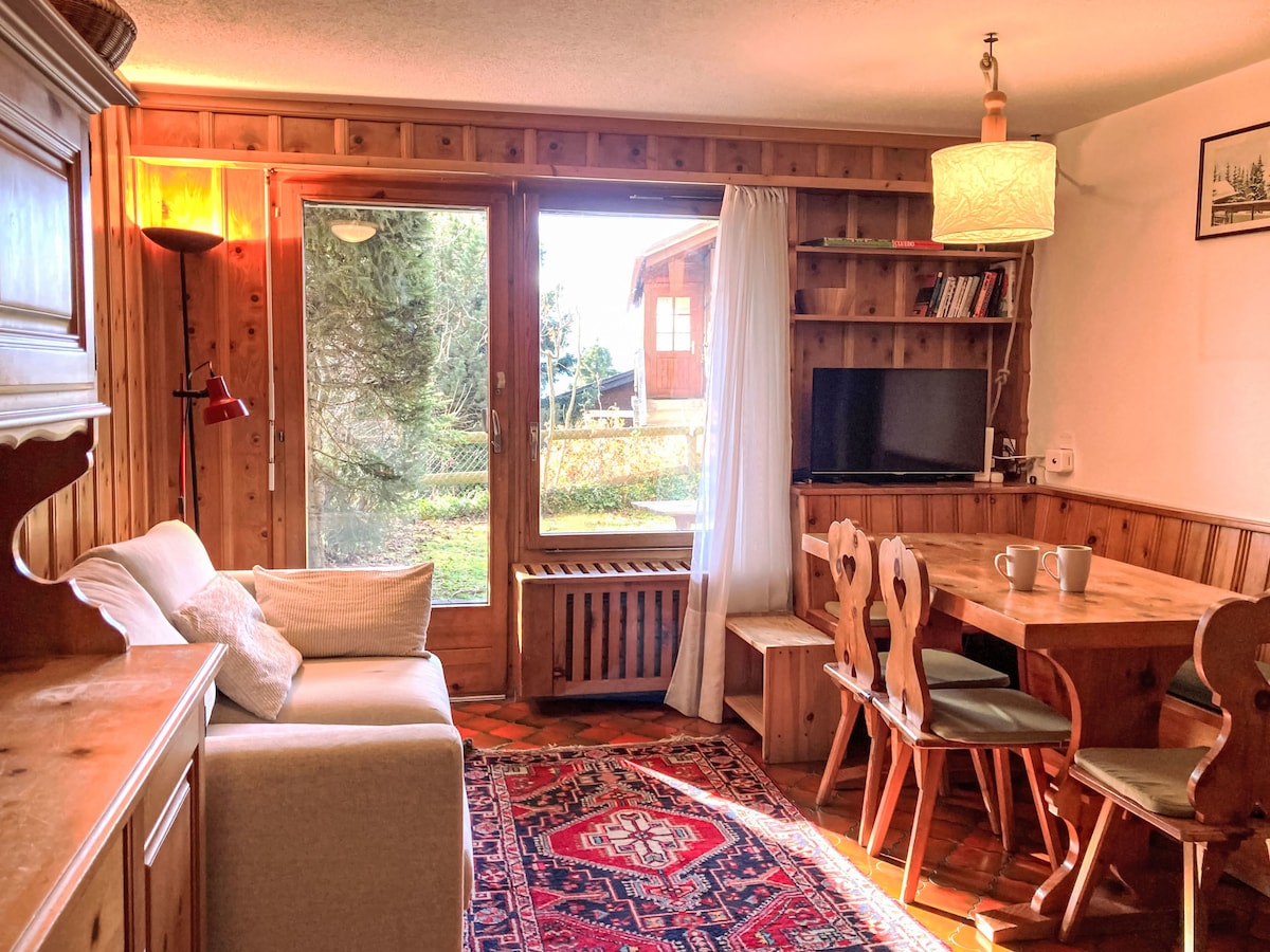 A cozy living and dining area is showcased, featuring a wooden table with seating for four and a small TV on a shelf. Natural light enters through large windows, illuminating the comfortable sofa and warm wood accents throughout the space.