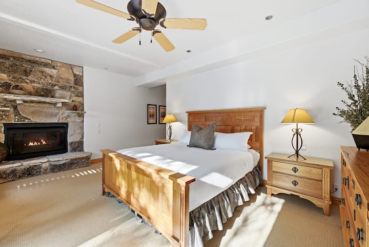 Dormitor confortabil cu șemineu din piatră, ventilator de tavan și mobilier din lemn în tonuri calde, inclusiv o tăblie și noptiere impunătoare.