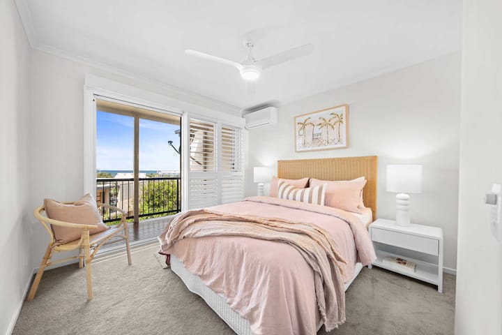 Une chambre au style côtier chic avec un lit queen size, des aperçus sur l'océan et un accès au balcon partagé avec la chambre principale.