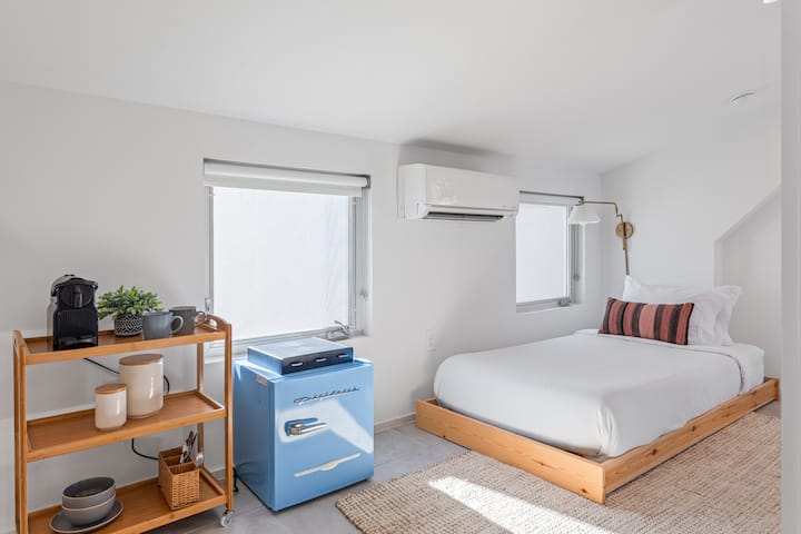 Mwonekano wa studio unaonyesha kitanda, friji ndogo ya bluu na rafu.