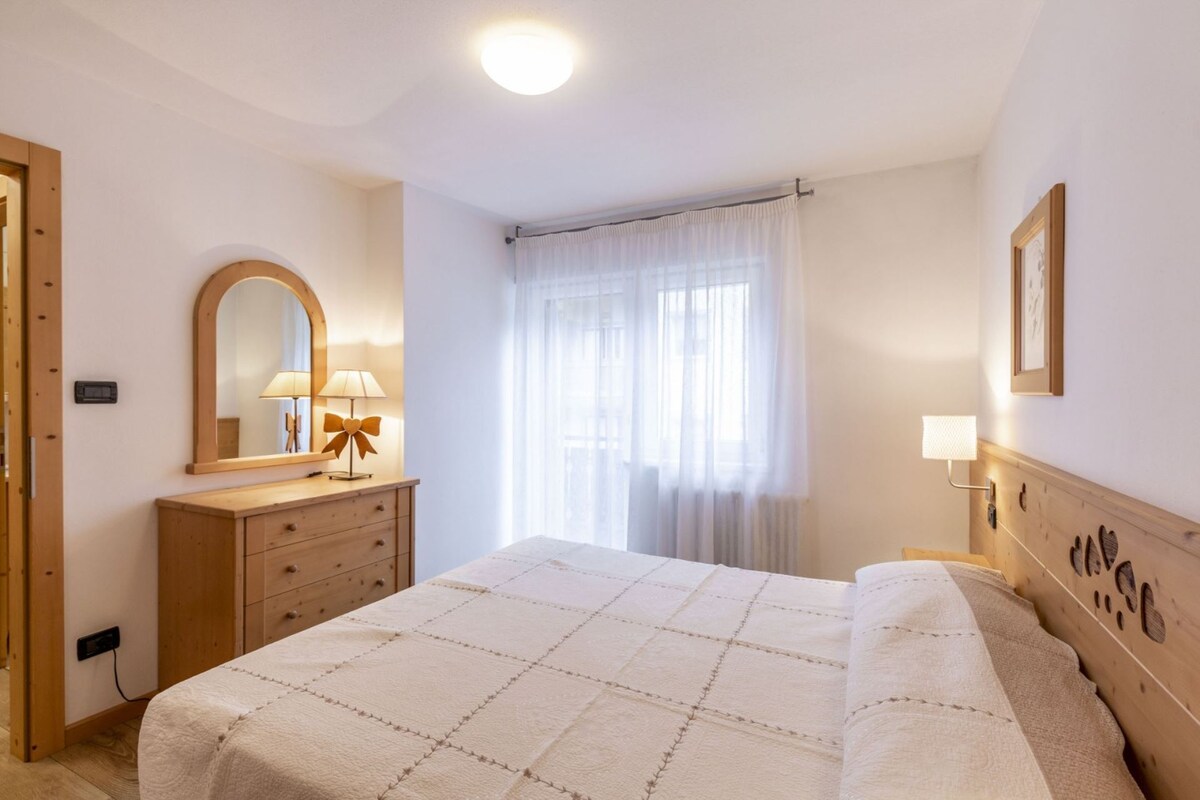 A comfortable double bedroom features a spacious bed adorned with a neutral-toned blanket. Natural light fills the room through sheer curtains. A wooden dresser and mirror complement the warm decor, while soft lighting from nightstands enhances the cozy ambiance.