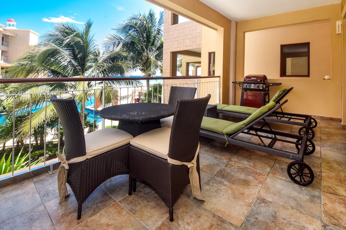 The private terrace is furnished with a round dining table and four wicker chairs. Two lounge chairs are positioned nearby, offering a view of the pool and surrounding palm trees. Light tile flooring complements the outdoor space, with natural light enhancing the atmosphere.