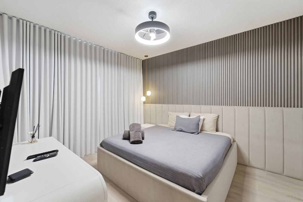 Disfruta de un acogedor dormitorio con una elegante cama tapizada con sábanas de color gris suave, realzada por paneles de pared texturizados. La luz cálida de un accesorio de techo moderno crea un ambiente sereno, perfecto para noches de descanso.