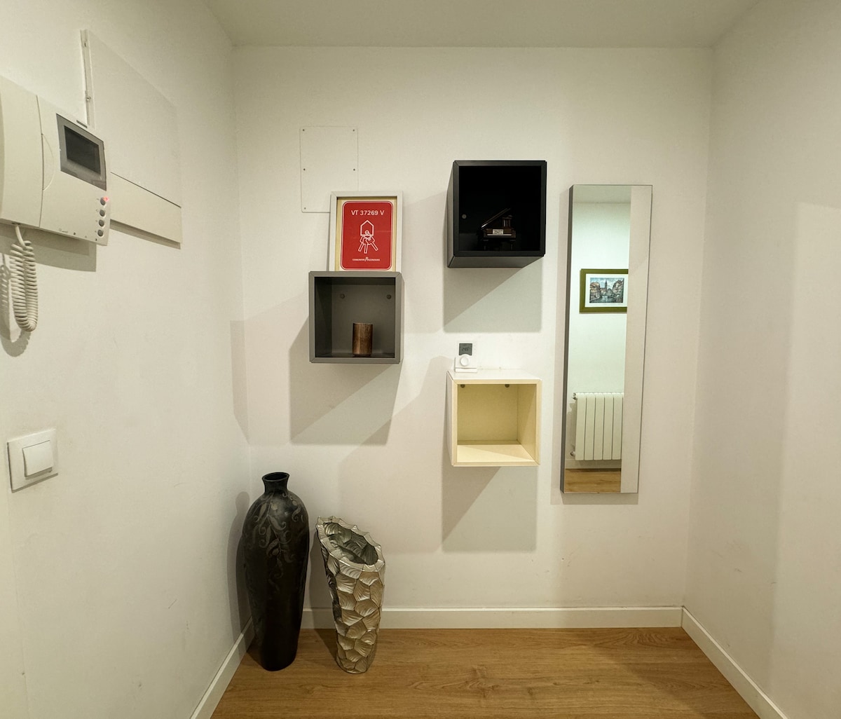 A minimalist entryway features a variety of wall-mounted boxes in black and white, displaying decorative items. A large dark vase and a light-colored sculptural vase are placed on the wooden floor, while a full-length mirror reflects the spaciousness of the area.