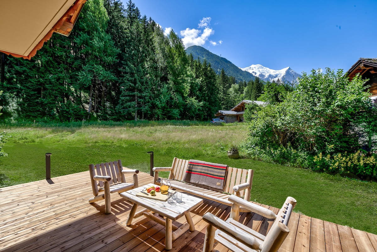 A wooden deck is furnished with comfortable seating, inviting relaxation in the fresh air. A small table is set with refreshments. The surrounding landscape features lush greenery, while mountains can be seen in the background under a clear blue sky.