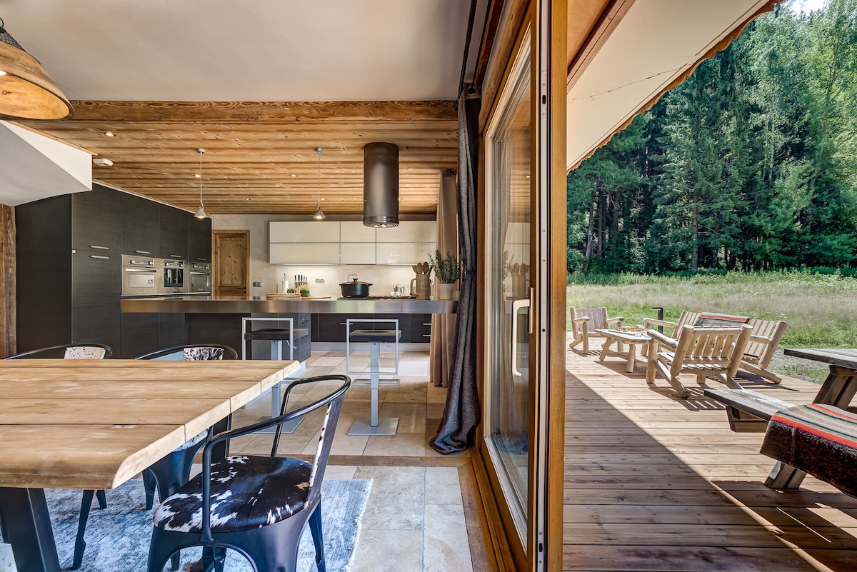An open-plan dining area is visible, featuring a wooden table surrounded by modern black chairs. The adjacent kitchen area has sleek cabinetry and modern appliances. Large sliding glass doors lead to an outdoor deck with wooden seating, surrounded by greenery, enhancing the connection to nature.