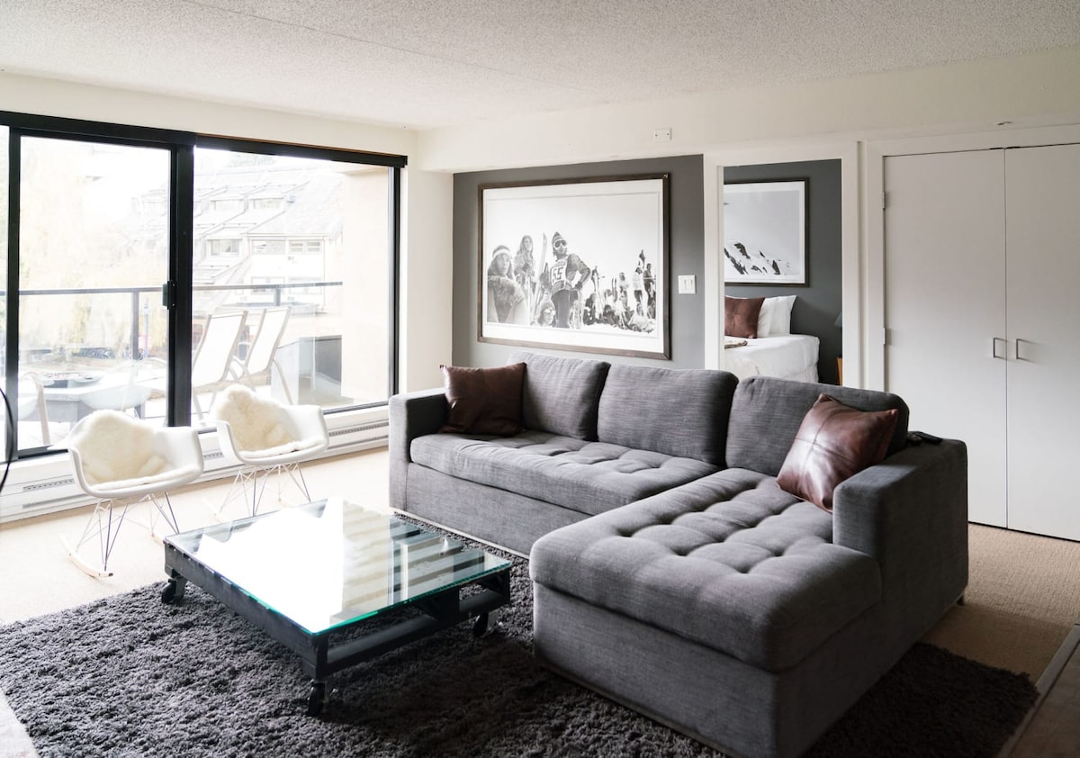 A modern living area features a spacious gray sectional sofa with textured pillows, positioned beside a large glass coffee table. Large windows provide natural light and views of the outdoor balcony. A framed black-and-white photograph adorns the wall, enhancing the contemporary feel.