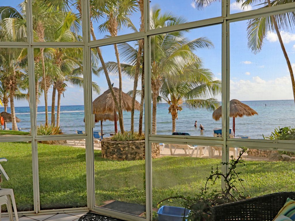 A view from inside showcases a screened patio overlooking the beach. Lush green grass stretches towards the sparkling water, with palm trees swaying gently. Thatched umbrellas provide shade on a sandy area where people can be seen enjoying the ocean.
