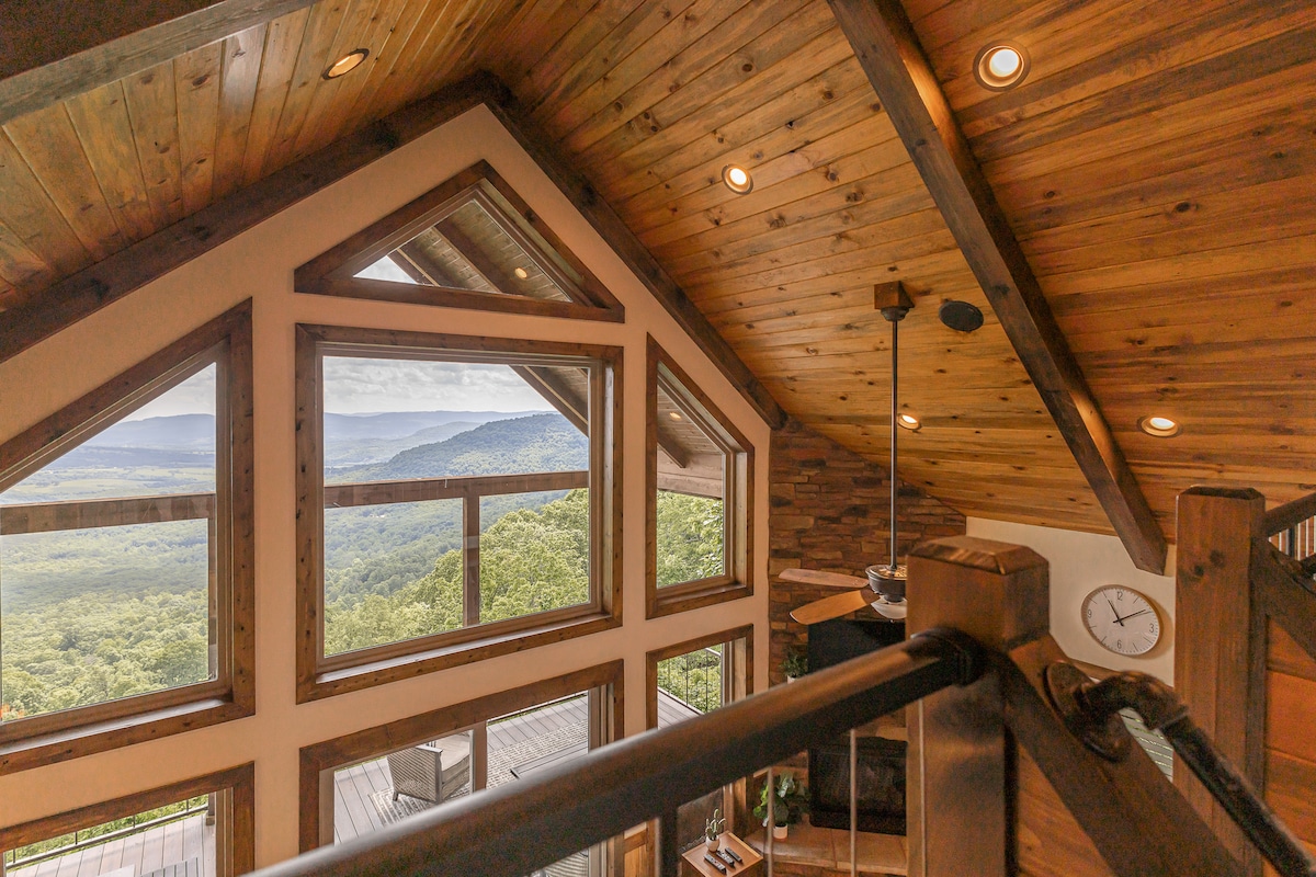 Large windows frame expansive views of the Arkansas Grand Canyon, inviting natural light into the open loft space. The wooden beams and ceiling add warmth to the design, while a stone fireplace anchors the room. A clock is mounted on the wall, emphasizing the serene atmosphere.