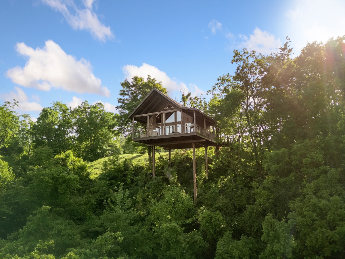 A treehouse perched among lush greenery is presented, featuring a large balcony that offers views of the surrounding landscape. The structure is elevated above the ground, supported by wooden beams, set against a backdrop of blue skies and scattered clouds.