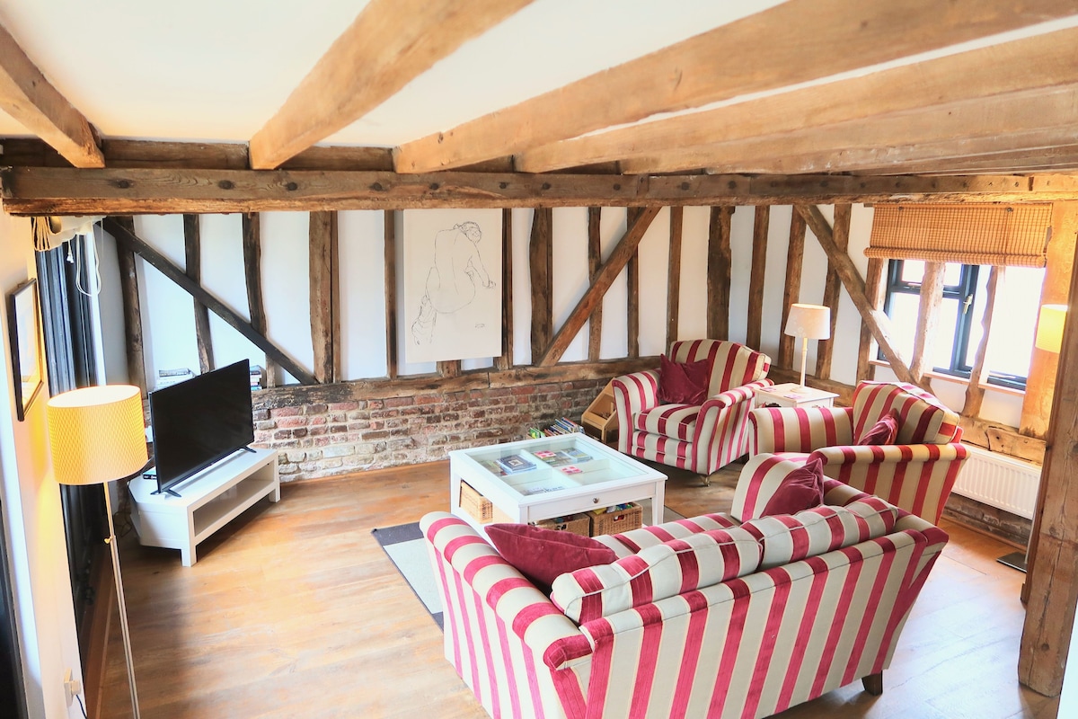 An open living space is showcased with exposed wooden beams and a combination of brick and white walls. Two striped couches face a coffee table in the center, while a flat-screen television is positioned on a stand. Soft light is provided by nearby lamps.