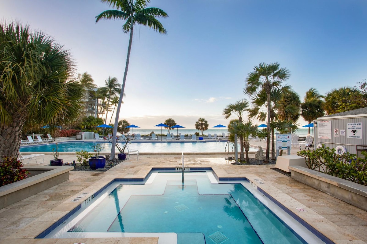 A heated whirlpool with clear water is positioned in the foreground, surrounded by lush landscaping and palm trees. In the background, a large heated pool and beach umbrellas create a serene atmosphere, offering a view of the Atlantic Ocean.