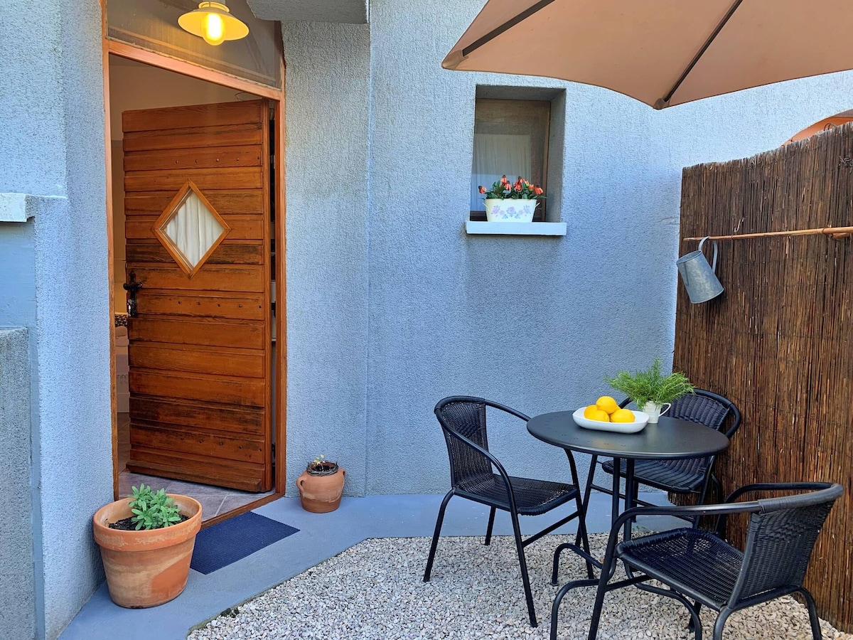 A welcoming entrance is highlighted by a wooden door with a diamond window, surrounded by soft gray walls. A round table with four black chairs sits on a pebbled surface, adorned with a bowl of fresh lemons and small potted plants. A large umbrella provides shade.