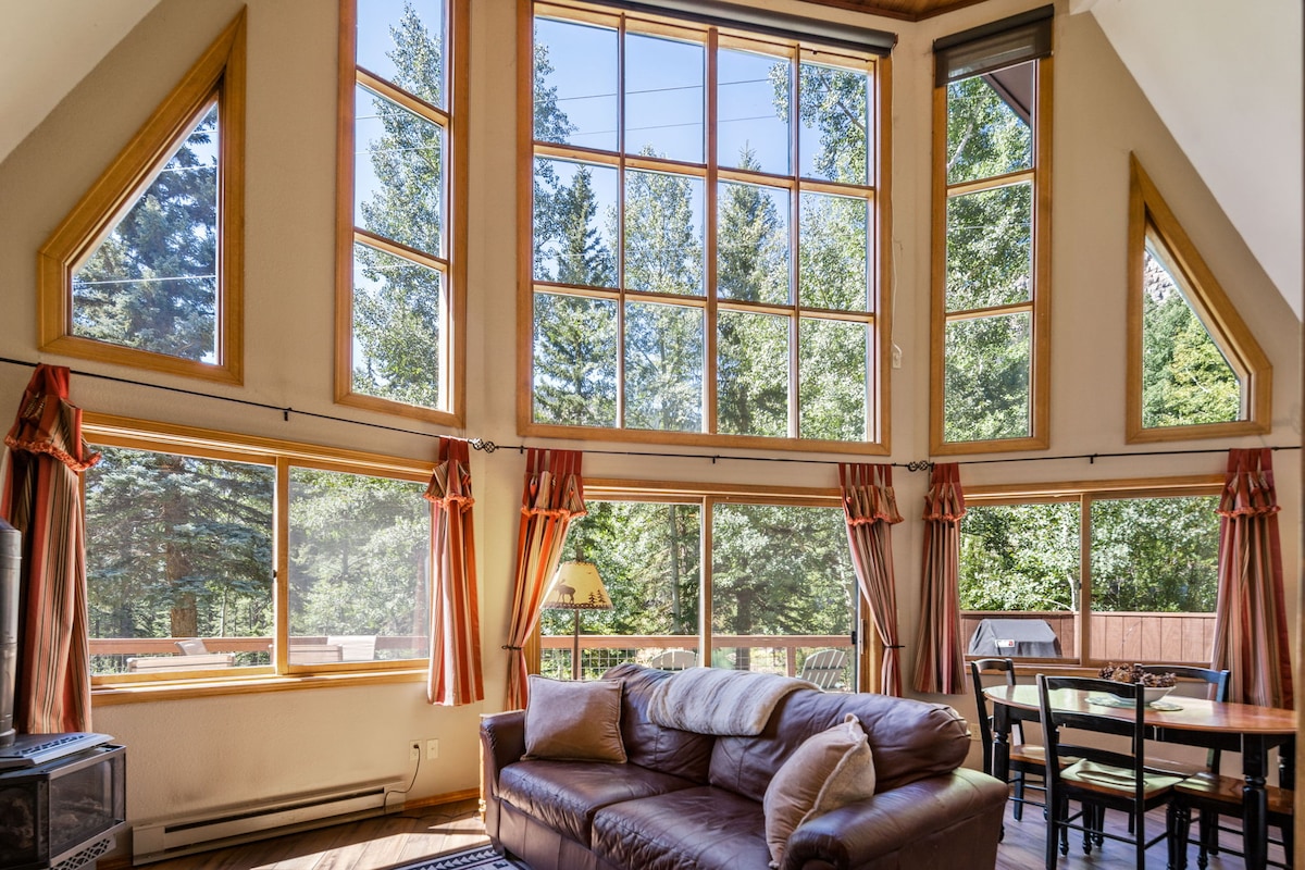 A spacious living area is highlighted by expansive windows that offer views of the surrounding trees. Natural light illuminates the room, which features a comfortable sofa and a dining table set for four. A wood-burning stove adds a cozy element to the space.