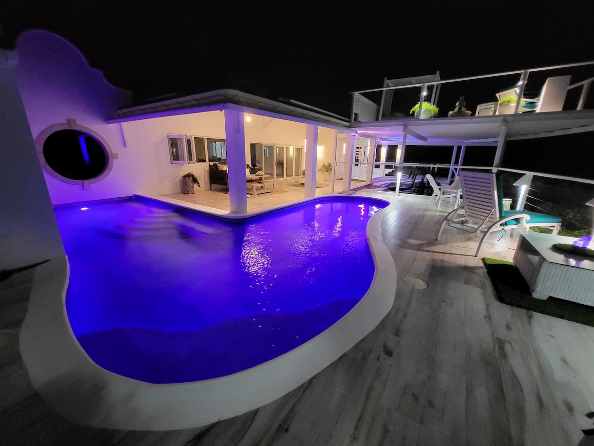 The villa's wraparound pool is illuminated in shades of blue, creating a serene night ambiance. Multiple seating areas can be seen on the upper deck, offering relaxed spaces for socializing. Soft lighting enhances the contemporary architecture, highlighting the open design and inviting atmosphere.