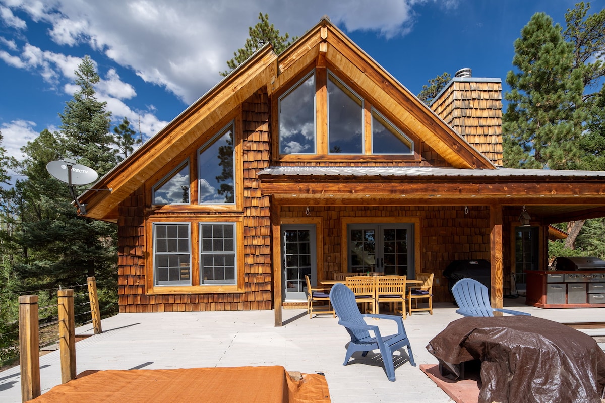 The exterior of the cabin features natural wooden shingles and large windows allowing for ample light. A spacious deck is shown, furnished with wooden seating and a cozy area for relaxation. Surrounding pine trees complement the serene mountain atmosphere.