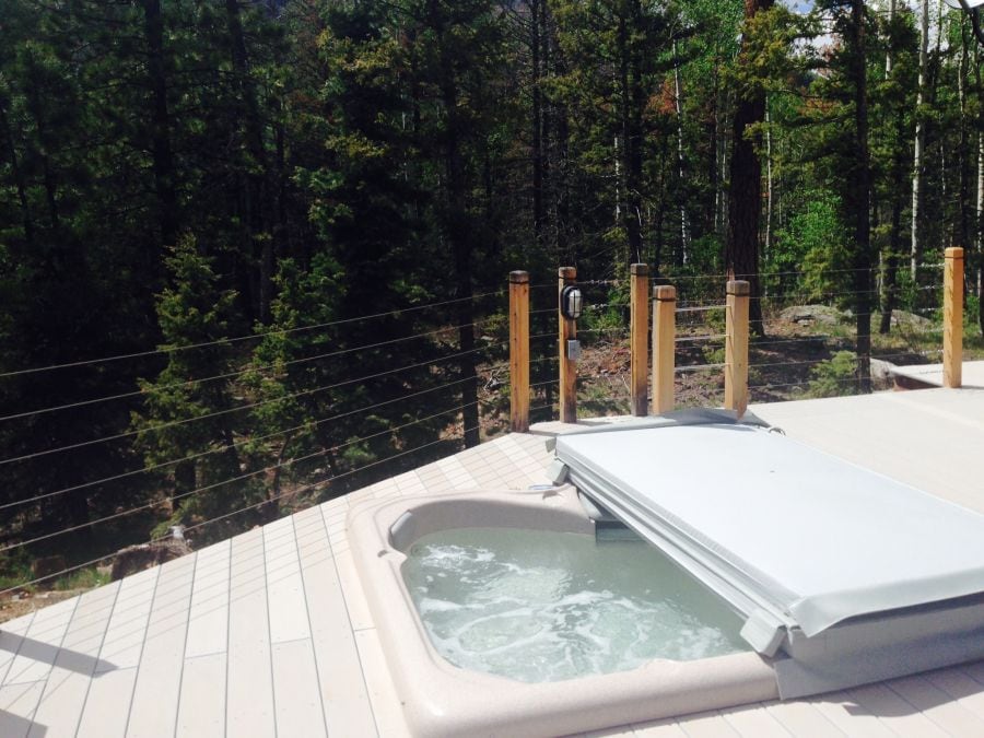 A private jacuzzi is positioned on a spacious deck, surrounded by tall trees and natural landscape. The tranquil water is shown bubbling gently, with a protective cover resting nearby. Glass railings offer unobstructed views of the serene forest setting.