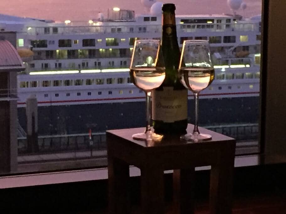 A cozy scene features a small wooden table holding two wine glasses filled with sparkling wine, accompanied by an unopened bottle. In the background, a large cruise ship is visible through the window, illuminated by the soft light of dusk.