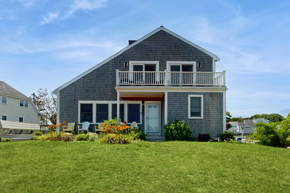 The exterior of the beach house features a well-maintained grassy front yard with vibrant flower gardens. A welcoming porch with seating is visible, framed by large windows. The structure showcases a distinctive gray shingle design and a second-floor balcony, enhancing the coastal charm.