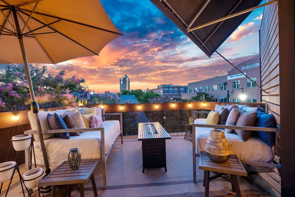 An outdoor terrace is showcased, featuring day beds adorned with cushions, and a coffee table in the center. An umbrella provides shade, while twinkling lights line the railing. The vibrant sunset paints the sky in warm hues, enhancing the inviting ambiance of the space.