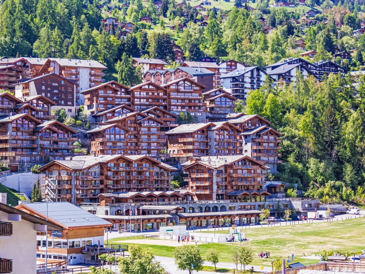 A scenic view captures a series of multi-level mountain residences, designed with wooden accents and balconies. The surrounding greenery provides a natural backdrop, while an open field in the foreground offers space for recreation. The image illustrates a vibrant alpine community.