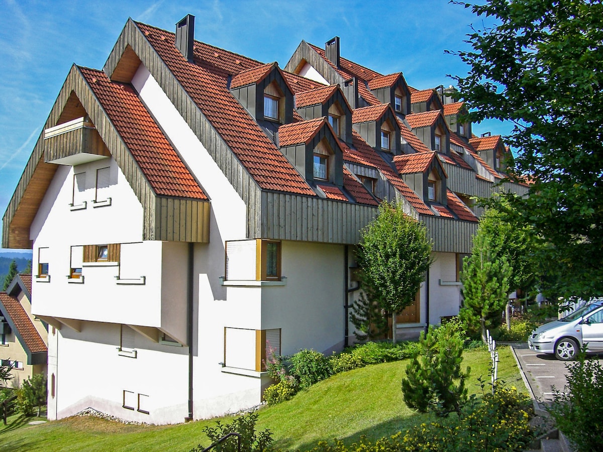 The exterior of the resort showcases an architectural design with multiple triangular roofs, complemented by a combination of wooden and white facades. Lush greenery and well-maintained landscaping surround the structure, providing a serene environment. A parking area is visible nearby.