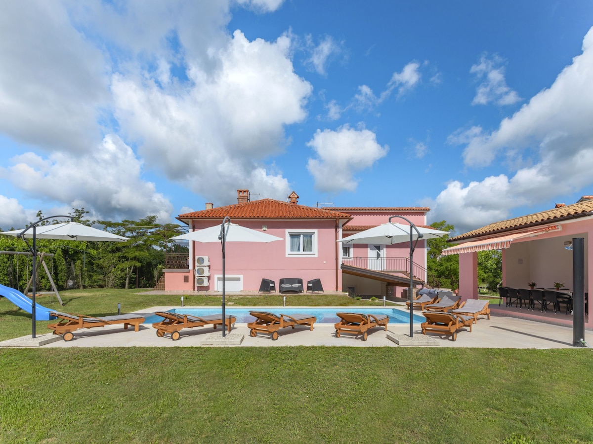 A spacious outdoor area features a swimming pool surrounded by sun loungers. A covered terrace with outdoor dining furniture and umbrellas provides shade. The house, with a pink exterior, is visible in the background against a bright blue sky with clouds.