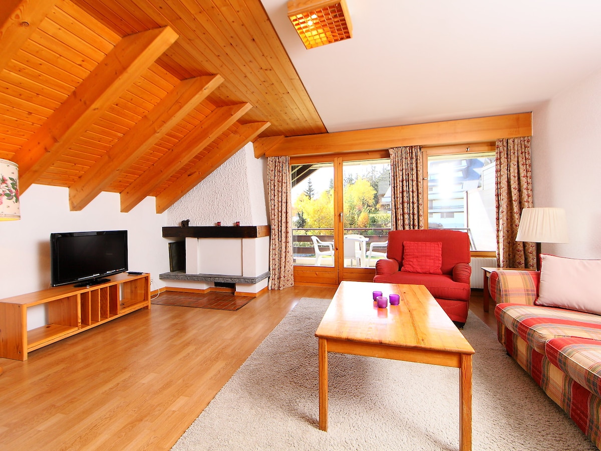 The living/dining room features sloping wooden ceilings and warm wooden flooring. A flat-screen TV is placed on a wooden console, while a cozy red armchair and a patterned sofa sit near a large window that opens to the balcony.