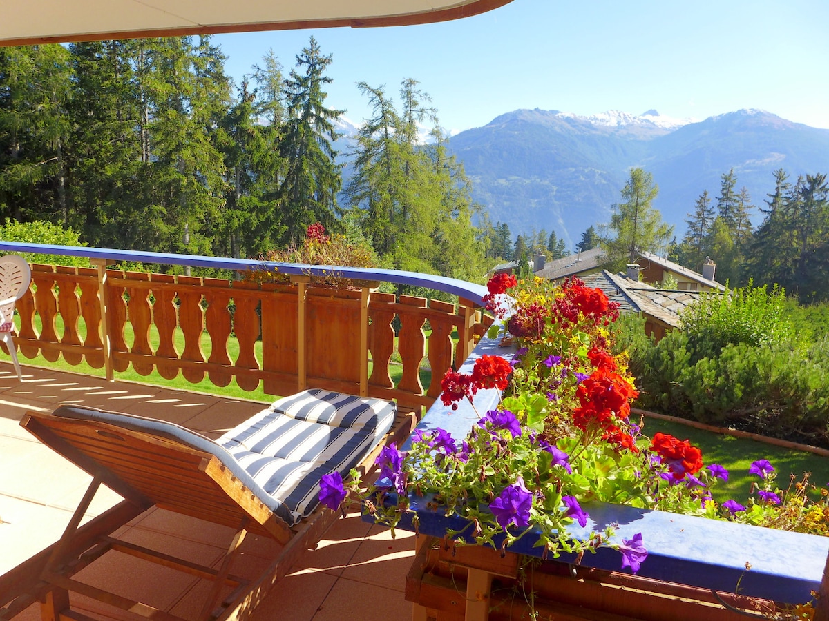 A large balcony is bordered by vibrant flower arrangements in red and purple. A reclining chair is positioned for relaxation, offering a view of snow-capped mountains and lush trees in the distance. The area is bathed in natural light.