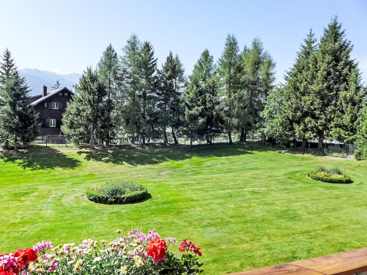 A well-maintained garden is featured, showcasing a spacious lawn that is bordered by tall trees. In the foreground, colorful flowers add vibrancy to the view. The serene landscape provides a tranquil atmosphere, highlighted by the distant outline of the building against a clear sky.