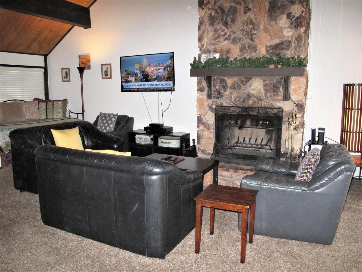 A comfortable living area is showcased with dark leather sofas arranged around a wooden coffee table. A stone fireplace serves as a focal point, complemented by a flat-screen TV mounted above. Soft lighting is provided by a stylish lamp, enhancing the relaxed atmosphere.