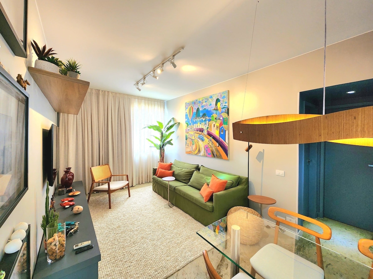 A modern living area features a green sectional sofa and a wooden chair, complemented by a large piece of colorful wall art. Natural light filters through sheer curtains, illuminating a neutral-toned space with decorative plants and contemporary furnishings.