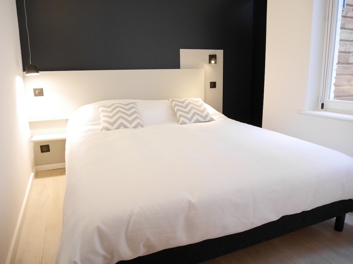 A spacious bed is presented with a crisp white duvet and two patterned pillows. The wall behind the bed features a contrasting dark panel, while softly-lit wall sconces provide gentle illumination. A window allows natural light to enhance the room's minimalist aesthetic.