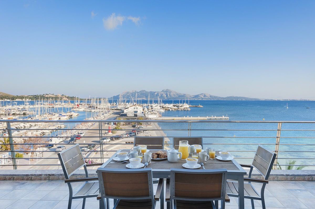 A dining area on a spacious terrace overlooks the marina and sea. A table is set with breakfast items, including fruit, pastries, and beverages, while the background features sailboats and a picturesque shoreline under a clear blue sky.