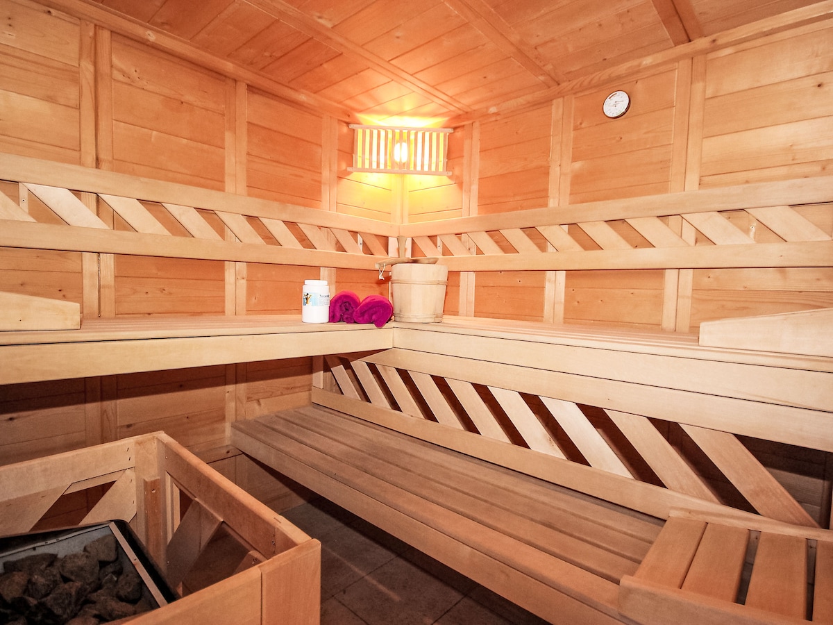 The interior of a wooden sauna features built-in seating along the walls and a stone heater in the corner. A soft glow is emitted from a light fixture mounted on the wall, enhancing the cozy atmosphere. Towels are neatly arranged on the bench.