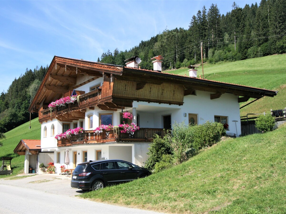The country house features a traditional Alpine design, with a wooden balcony adorned with vibrant flowers. A parked car is visible in the foreground, while green hills and trees create a serene backdrop, enhancing the peaceful setting.