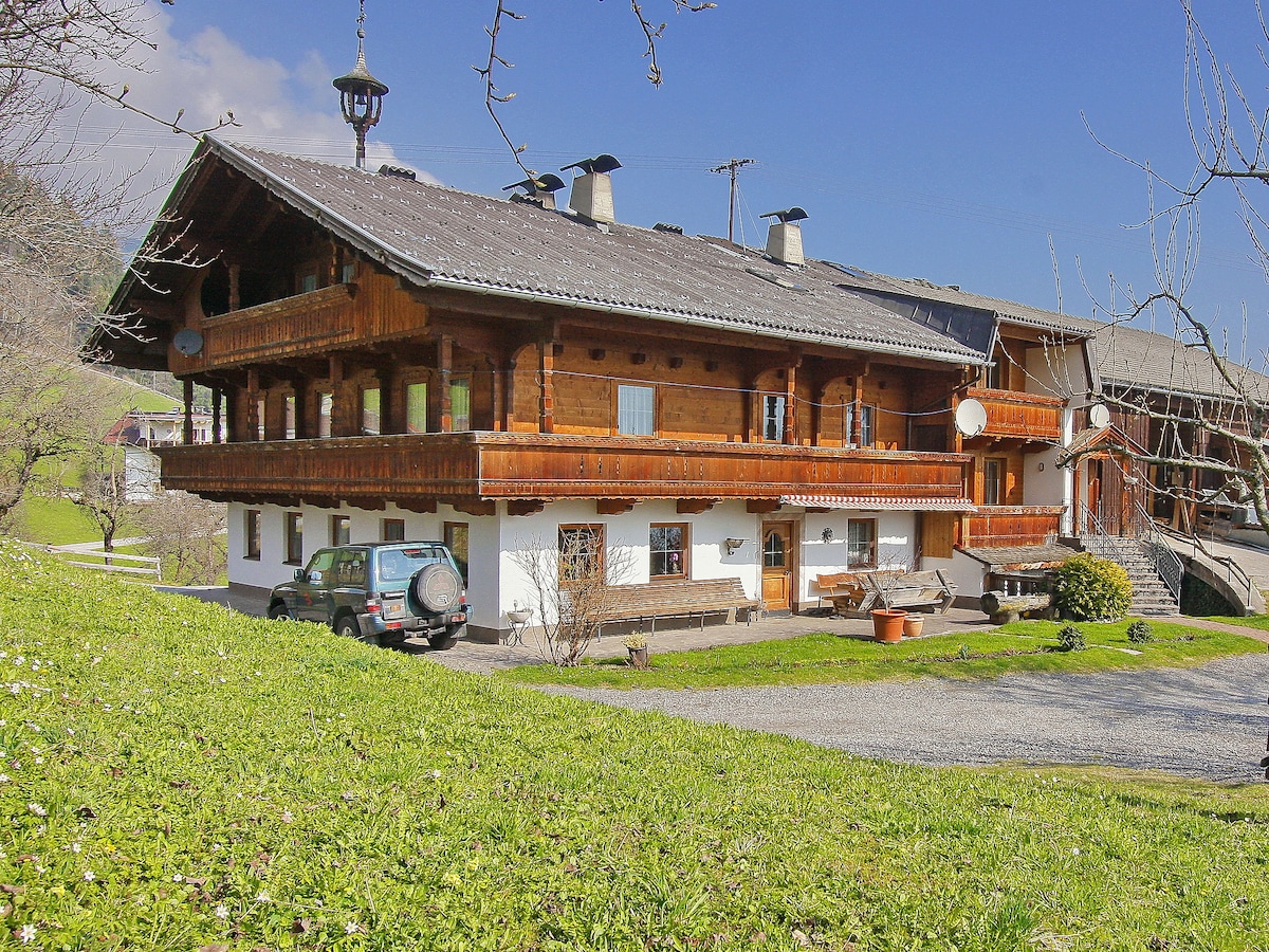 A multi-level wooden structure is set amidst a grassy area, featuring a large balcony with traditional wooden railings. The facade includes several windows, and satellite dishes are mounted on the exterior. A vehicle is parked nearby on a gravel surface.