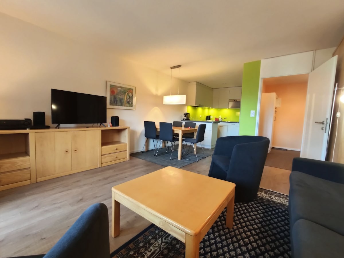 A brightly lit living and dining area features modern furnishings, including a flat-screen TV and a wooden dining table with six chairs. Light green walls accent the open kitchen equipped with contemporary appliances. A cozy seating arrangement with dark chairs and a light wooden coffee table enhances the space.