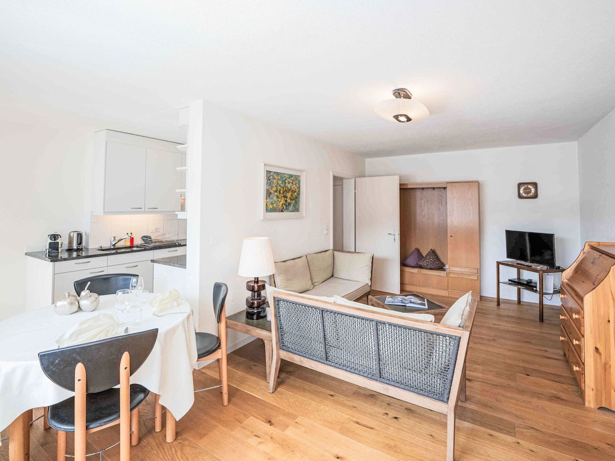 The living/dining area features a light-colored sofa paired with wooden furniture, including a dining table set for four. A flat-screen TV is positioned against the wall. Warm wood flooring complements the open kitchen, which includes modern appliances and a window providing natural light.