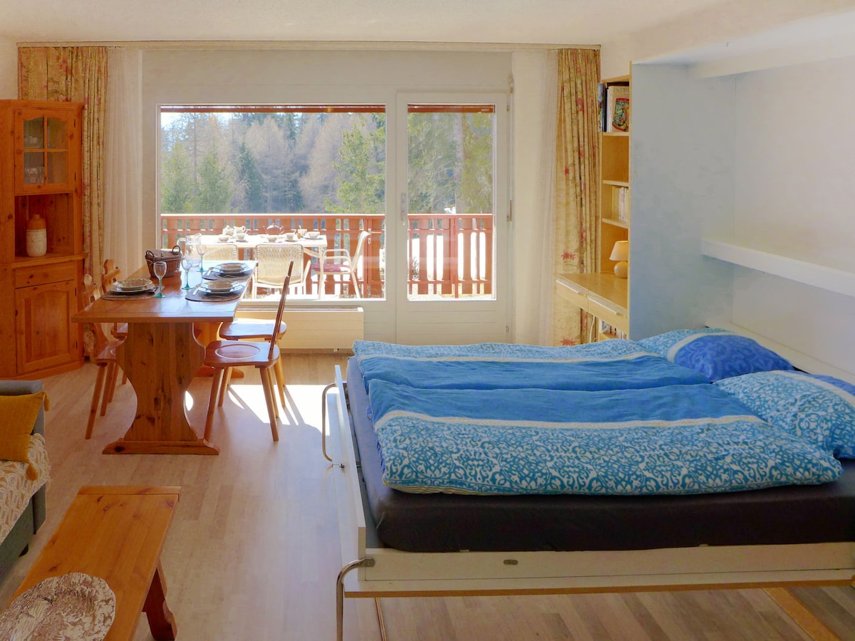 A spacious living area is shown with a sofa, a dining table set for four, and a cozy bed spread with a blue blanket. Large windows offer a view of the forest outside and provide natural light to the room.