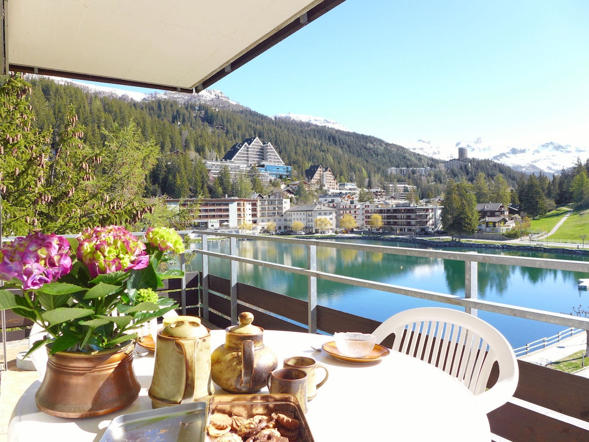 A balcony table is set with a flower arrangement, a teapot, and plates of cookies. The view features a serene lake bordered by greenery and mountains, with a sunny sky in the background. Scenic buildings are visible along the lakefront.