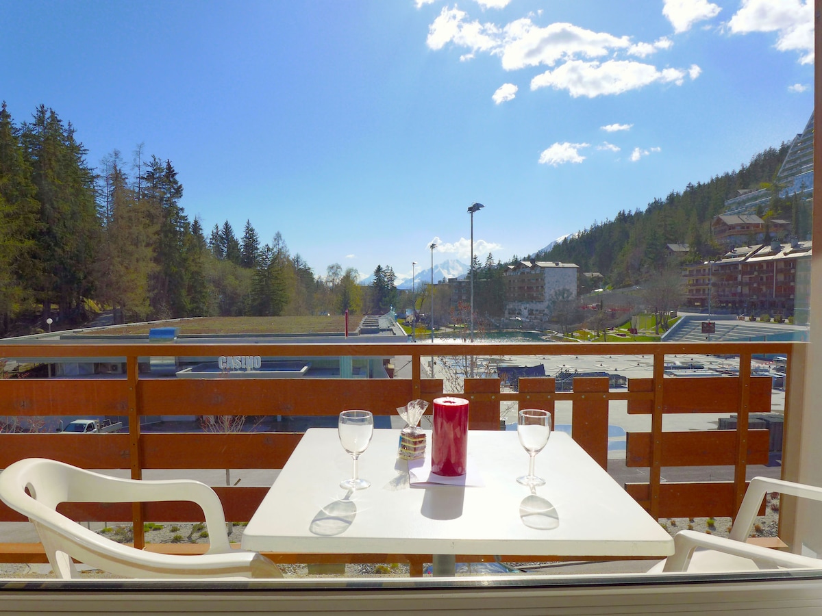A balcony features a white table set for two, with two glasses positioned on it. The view includes distant mountains and greenery, alongside a glimpse of neighboring buildings under a clear blue sky.