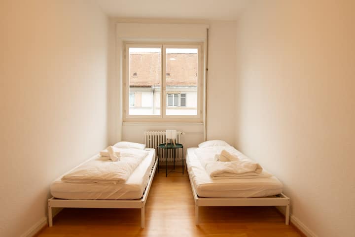 Un dormitorio tranquilo y aireado con amplia luz natural, que ofrece dos camas individuales en un espacio limpio y despejado.