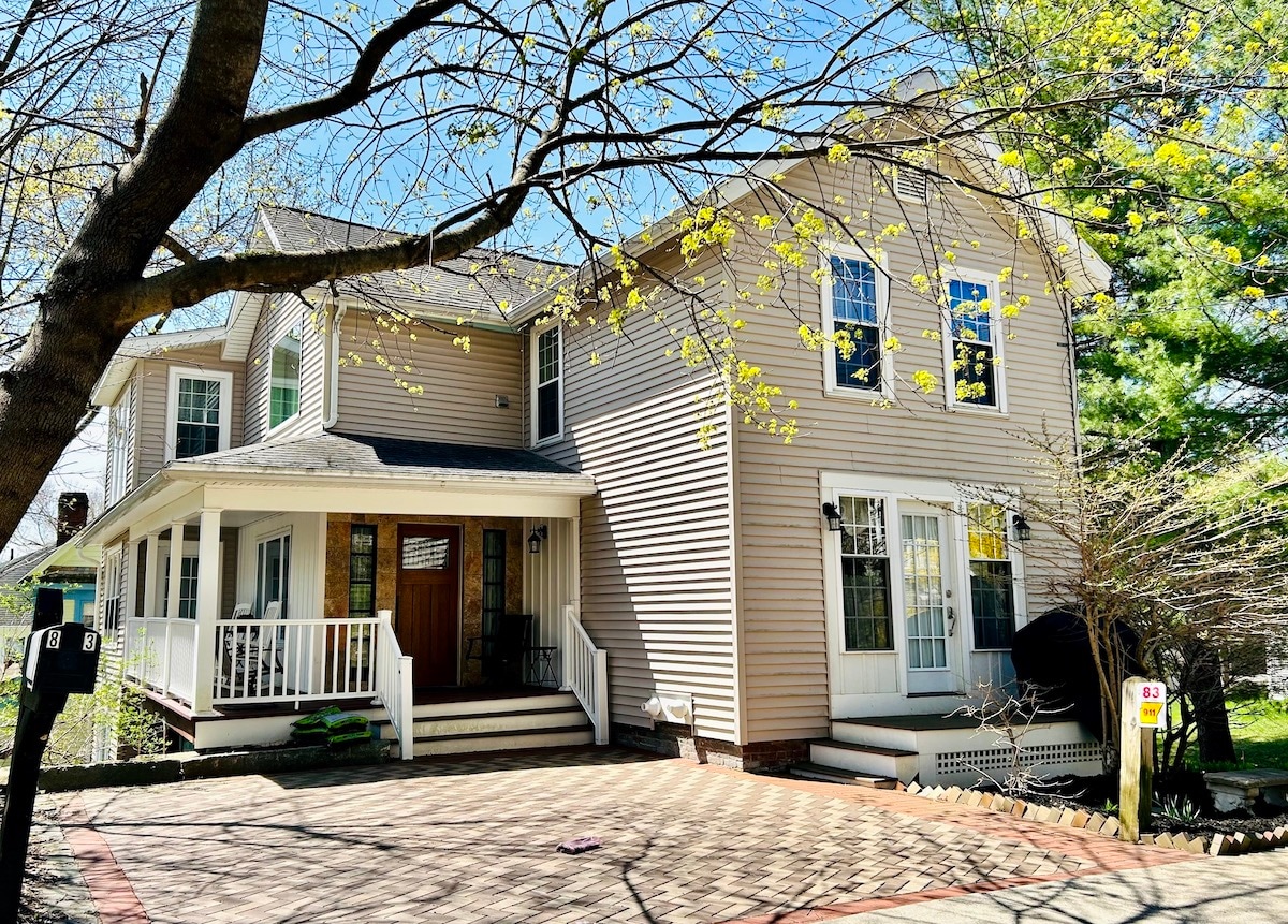 The exterior of a two-story townhouse is shown, featuring a welcoming front porch with white railings. Large windows allow natural light to fill the space. A paved driveway leads up to the entrance, surrounded by blooming trees, indicating a serene residential setting.