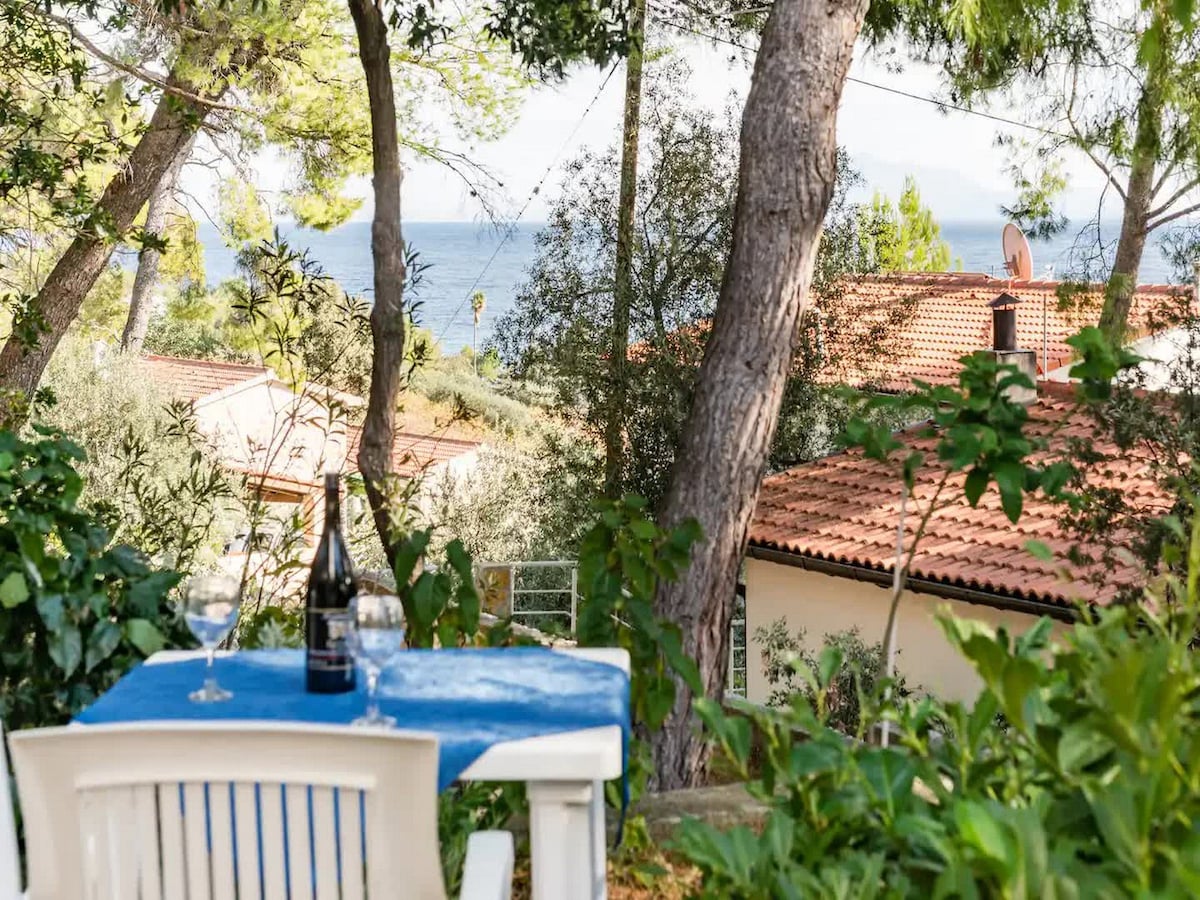 A table with a blue tablecloth set for two is positioned among greenery, offering views of nearby rooftops and the glistening coastline. A bottle and two glasses are visible, enhancing the inviting outdoor dining experience surrounded by trees and lush foliage.