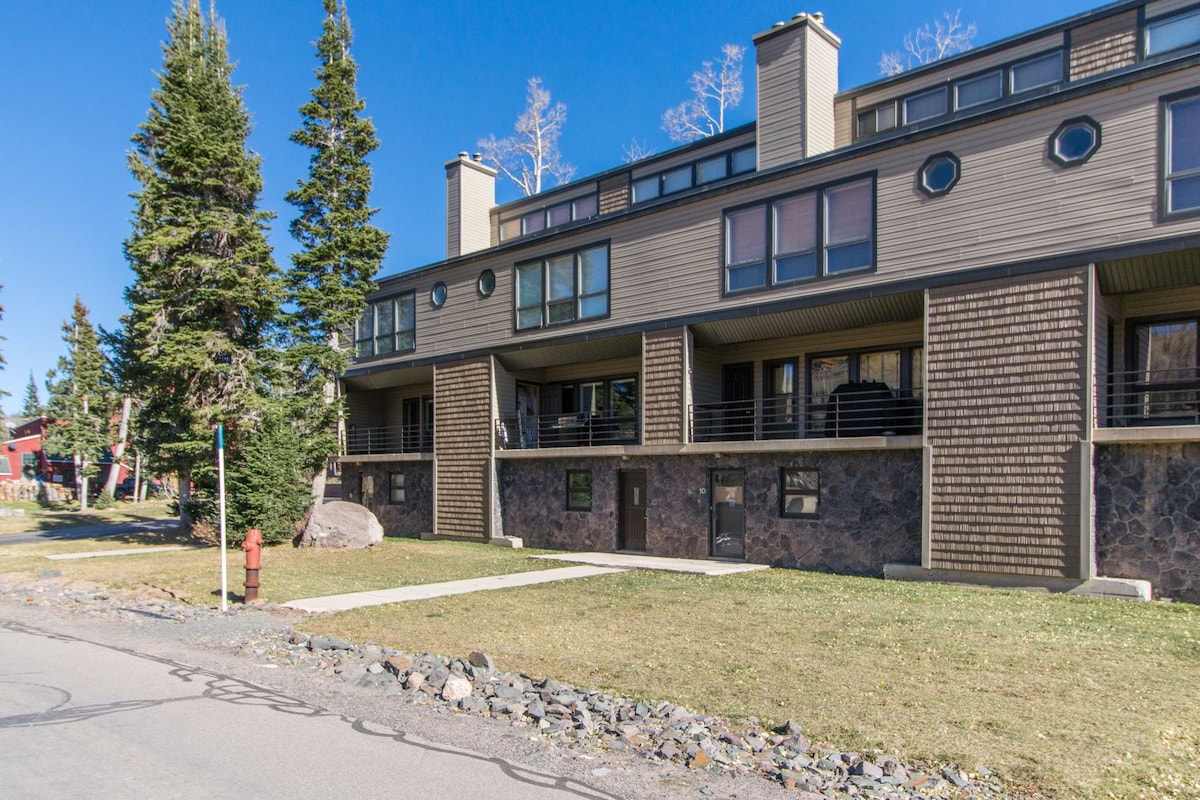 The exterior of a multi-level condo is showcased, featuring a combination of wood and stone elements. Large windows provide natural light, while a grassy area with a path is visible in front. Surrounding trees contribute to the natural setting.