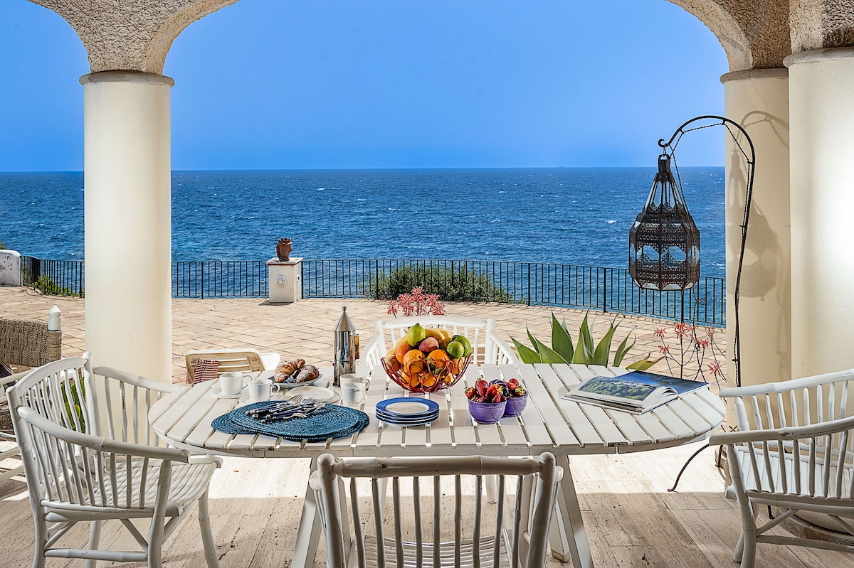 An outdoor dining area features a long table set with colorful dishes of fresh fruit. The sea view is framed by elegant columns, and a lantern hangs beside the table, adding to the serene coastal ambiance.