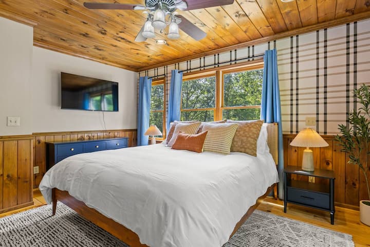 Unwind in this peaceful bedroom featuring a comfortable king bed, warm wood accents, and large windows with tranquil forest views. Enjoy cozy lighting, soft bedding, and a mounted TV for relaxing after a day of adventure.